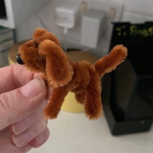 Op de afbeelding: Een bruin pijpenrager hondenbeeldje met een zachte textuur. De hond heeft slappe oren, een kleine staart en zwarte kraalogen. Het handgemaakte ambacht wordt in een hand gehouden.