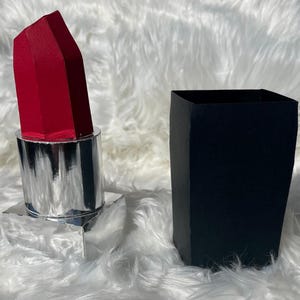 May include: A three-dimensional paper craft model of a lipstick tube. The lipstick features a bright red, faceted bullet, a reflective silver metallic base, and a separate, solid black rectangular cap standing upright next to the main tube.