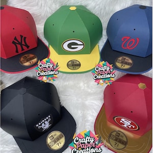 Peut inclure: Plusieurs casquettes de baseball en papier aux couleurs d'équipes, dont le rouge, le vert, le bleu et le noir. Chaque casquette affiche un logo d'équipe et le texte "59FIFTY 71". Les casquettes sont présentées sur une surface blanche et moelleuse, avec le texte "Crafty Paper Creations".