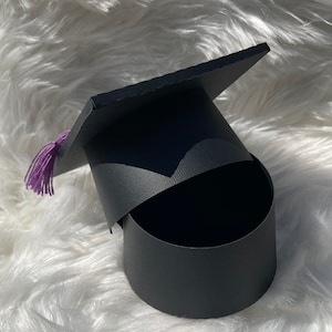 May include: Black graduation cap shaped box with a purple tassel. The box is open and the inside is visible.