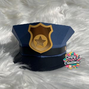Può includere: Un cappello da poliziotto di carta blu con un distintivo dorato a forma di stella. Il cappello ha un bordo nero e un distintivo dorato con una stella al centro. Le parole "Crafty Paper Creations" sono sul lato destro del cappello.
