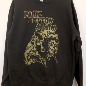 May include: A dark grey sweatshirt featuring the text "PANIC BUTTON AGAIN!" in a distressed font, above an olive green illustration of a face. The top has a crew neck and long sleeves.