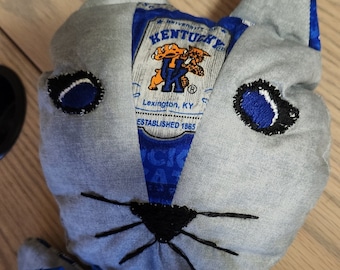 May include: A gray cat-shaped stuffed animal with blue and white embroidered eyes, a black embroidered nose and whiskers, and a blue and white patch on the chest that reads "University of Kentucky, Lexington, KY, Established 1865".