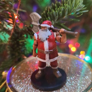 May include: Miniature figurine of Santa Claus, clad in a red and white suit, holding a large axe over his shoulder. The figure has a white beard and is posed on a black base. The background features a Christmas tree with colorful lights.