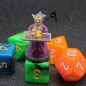 May include: A miniature figurine of a goblin server in a purple dress, holding a tray of baked goods. The figurine is placed on a green die, with other colorful dice in the background. The number "9" is written in the upper right corner.