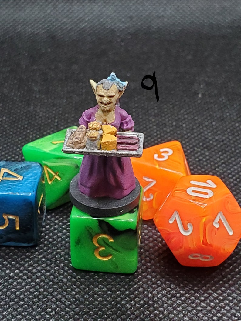 May include: A miniature figurine of a goblin server in a purple dress, holding a tray of baked goods. The figurine is placed on a green die, with other colorful dice in the background. The number "9" is written in the upper right corner.