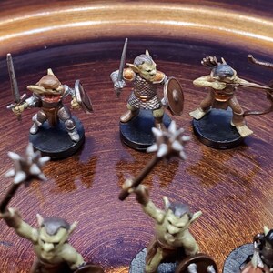 May include: Miniature fantasy figures of goblins and other creatures, painted in shades of brown, tan, and green. Some figures wield swords, shields, bows, and maces. The figures are on black circular bases, set against a brown background.