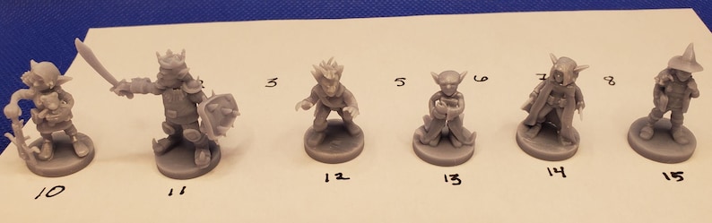 May include: A collection of gray miniature fantasy figures, each on a circular base. The figures include a warrior with a sword and shield, a wizard with a staff, and other characters. Numbers are written below each figure.