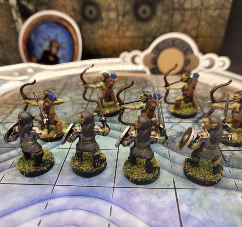 May include: Miniature fantasy figures of orcs, some with bows and arrows, others with swords and shields. The orcs are painted in shades of green, brown, and gray, standing on bases with grass. The figures are arranged on a blue and white grid.