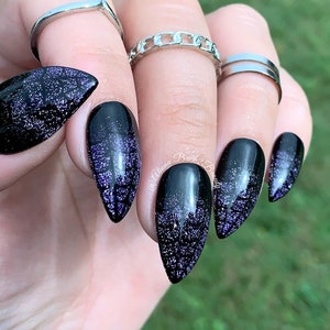 May include: Close-up of a hand with long, black, almond-shaped fingernails. The nails have a glittery purple gradient effect. Silver rings adorn the fingers. The background is blurred green.