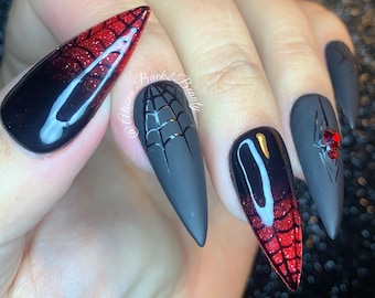 May include: Black stiletto nails with a spiderweb design and red glitter ombre. Some nails have a spiderweb design and a red rhinestone spider.