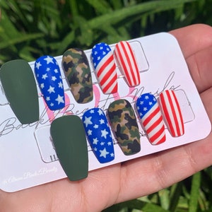 Puede incluir: Conjunto de diez u&ntilde;as postizas con tem&aacute;tica patri&oacute;tica. Las u&ntilde;as presentan una variedad de dise&ntilde;os, incluyendo camuflaje, estrellas y la bandera estadounidense. Las u&ntilde;as est&aacute;n en una tarjeta blanca con el texto "Back2Beauty" y "@iliana.back2.beauty".