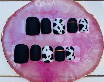 May include: A set of press-on nails in a variety of designs. Some nails are matte black, others feature a white and black cow print, and some have a copper-colored stripe. The nails are displayed on a pink and gold tray.