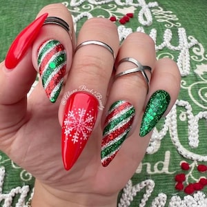 Op de afbeelding: Een hand met lange, amandelvormige nagels met kerstthema-ontwerpen. De nagels hebben rode, groene en zilveren glitterontwerpen, waaronder zuurstokstrepen en een sneeuwvlok. Zilveren ringen sieren de vingers.