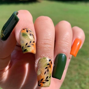 Op de afbeelding: Close-up van een hand met herfst-nagelkunst. De nagels zijn geverfd in tinten groen, oranje en goud, met een ontwerp van zwarte bladeren en gouden accenten. De nagelkunst heet "Autumn Back to Beauty".