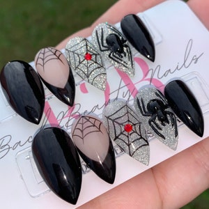 May include: Set of six black and silver glitter press-on nails with spider and web designs. The nails are in a stiletto shape and have a red rhinestone accent.