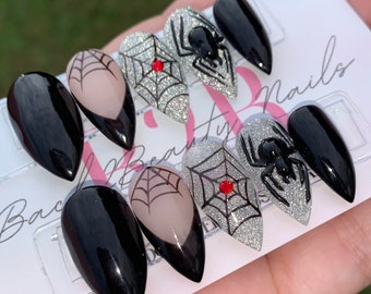May include: Set of six black and silver glitter press-on nails with spider and web designs. The nails are in a stiletto shape and have a red rhinestone accent.