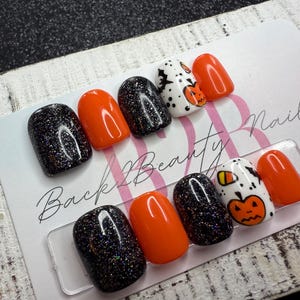 May include: A set of press-on nails with a Halloween theme. The nails feature a mix of black glitter, solid orange, and white with pumpkin and bat designs. The text "Back 2 Beauty Nails" is visible on the card.