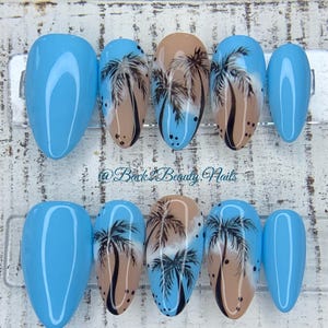 May include: Set of ten oval-shaped fake nails with a blue and beige base. Some nails have a palm tree design painted on them. The nails are arranged in two rows on a wooden surface. @Back2BeautyNails