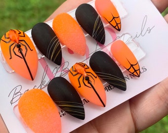 Op de afbeelding: Een set amandelvormige press-on nagels met een matte afwerking in zwart en feloranje. Sommige nagels hebben spinnenweb-lijnkunst, terwijl andere zijn versierd met een driedimensionale oranje kristallen spin op een oranje basis.