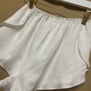 May include: White satin shorts with a ruffled edge and elastic waistband. The shorts are hanging on a white hanger.