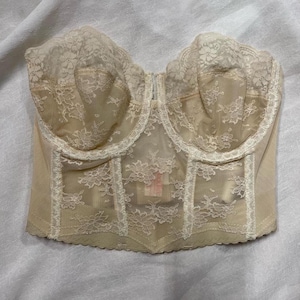May include: A beige lace strapless bra with a hook and eye closure in the back. The bra has a boned bodice and a floral lace pattern.