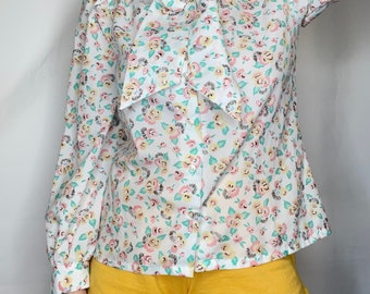 Puede incluir: Una blusa blanca de manga larga con estampado floral y un gran lazo de corbata en el cuello. La blusa tiene un frente abotonado y está hecha de una tela ligera.