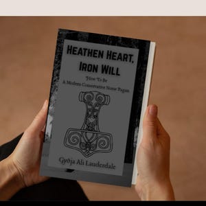 May include: A book titled "Heathen Heart, Iron Will" is held in hands. The cover features a stylized hammer design and the text "How To Be A Modern Conservative Norse Pagan" by Gyðja Ali Lauderdale.