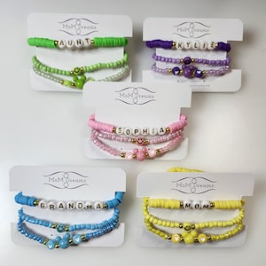 May include: Five handmade bracelets with the text "AUNT", "KYLIE", "SOPHIA", "GRANDMA", and "MOM". Each bracelet features colorful beads, smiley face charms, and heart-shaped accents. The bracelets are displayed on white cards with the "MoM Dreams" logo.
