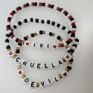 May include: Three beaded bracelets with black, white, red, and gold beads. One bracelet spells out "CRUELLA DEVIL" and another says "101". The bracelets are on a white surface.