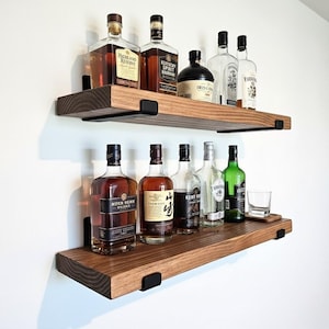 May include: Two wooden shelves with various liquor bottles, including whiskey and gin, are mounted on a white wall. The shelves are made of dark wood and supported by black metal brackets. A glass is on the bottom shelf.