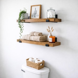 Set of two handcrafted rustic floating wood shelves in a bathroom above a toilet. These solid wood farmhouse shelves are styled with a potted plant, framed cactus art, and towels, featuring matte black metal J-brackets.