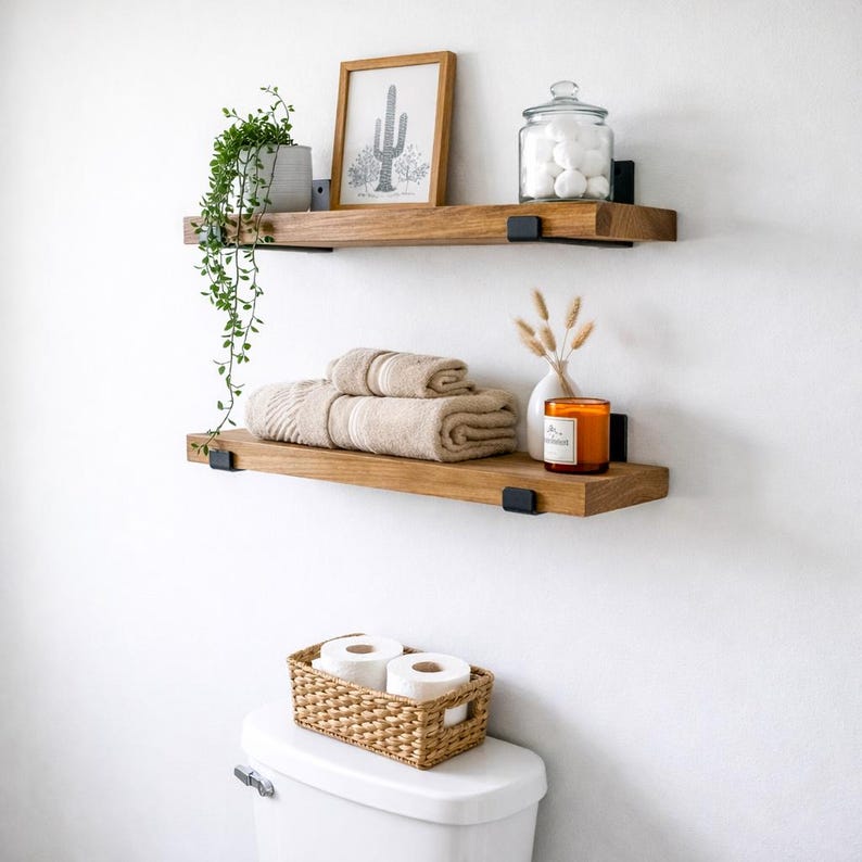 Set of two handcrafted rustic floating wood shelves in a bathroom above a toilet. These solid wood farmhouse shelves are styled with a potted plant, framed cactus art, and towels, featuring matte black metal J-brackets.