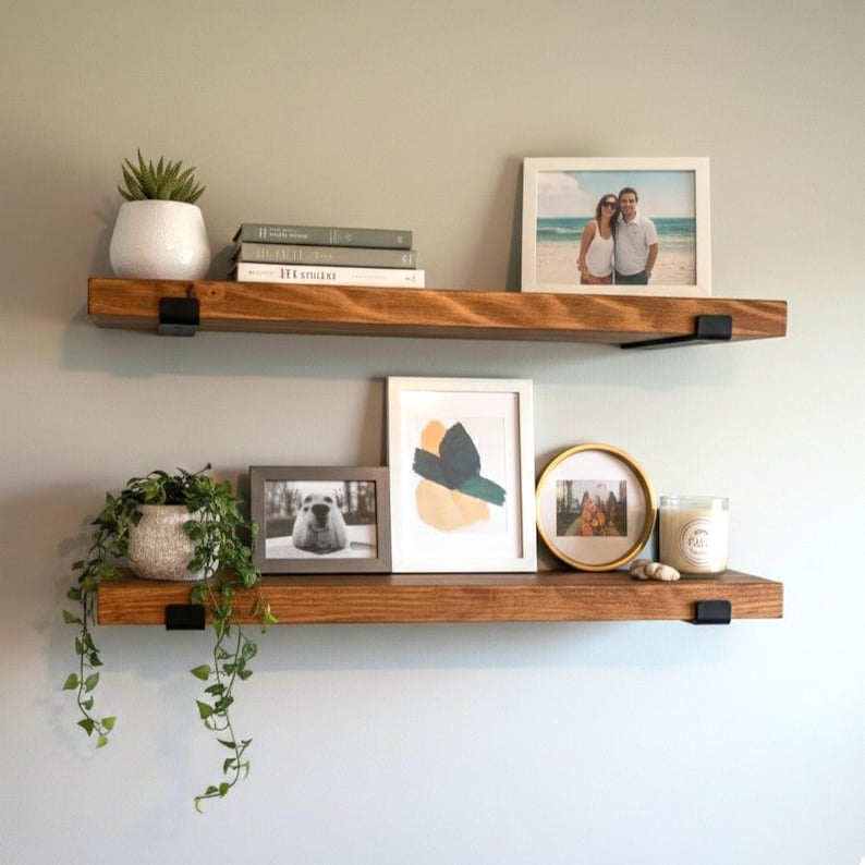 Peut inclure: Deux &eacute;tag&egrave;res flottantes en bois avec des supports en m&eacute;tal noir sont fix&eacute;es &agrave; un mur gris clair. Les &eacute;tag&egrave;res pr&eacute;sentent divers objets d&eacute;coratifs, notamment des photos encadr&eacute;es, des plantes en pot, des livres et une bougie.