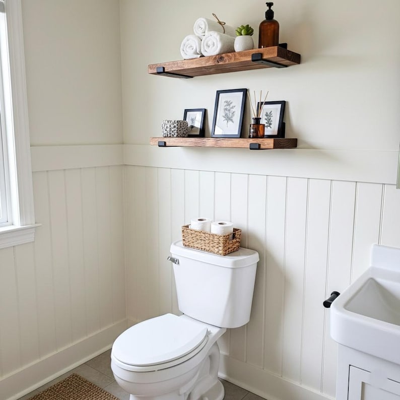 Peut inclure: Int&eacute;rieur de salle de bain avec murs blancs et lambris. Deux &eacute;tag&egrave;res en bois avec des supports m&eacute;talliques noirs supportent des serviettes blanches roul&eacute;es, une petite plante et des &oelig;uvres d'art encadr&eacute;es. Des toilettes avec un panier de papier toilette tress&eacute; sont en dessous.