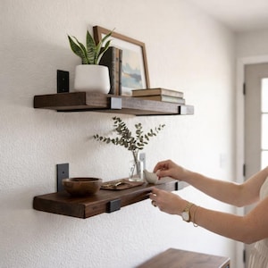 A person’s hands are seen placing a small ceramic bird on the bottom of two dark-stained wood floating shelves. The top shelf is decorated with a white potted plant and framed art, while the bottom shelf holds a wooden bowl and decorative accessories. Both shelves are supported by industrial black metal brackets against a neutral wall.