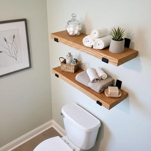 May include: Two wooden floating shelves with black brackets are mounted on a light green wall. The top shelf holds rolled white towels, a small potted plant, and a glass jar. The bottom shelf displays folded towels, a soap dish, and a basket.
