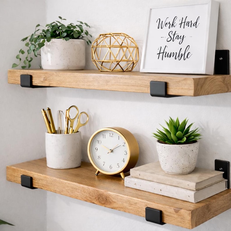 Set of two handcrafted rustic floating wood shelves in a bright home office. These solid wood shelves are styled with gold desk accessories, a modern clock, and a 'Work Hard Stay Humble' sign, held by matte black J-brackets.