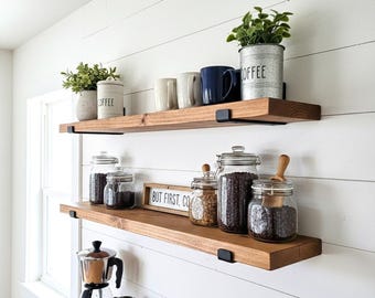 May include: Two wooden shelves with black metal brackets are mounted on a white wall. The top shelf holds mugs, a plant, and a container that says "COFFEE". The bottom shelf displays glass jars filled with coffee beans and a sign that reads "BUT FIRST, COFFEE".