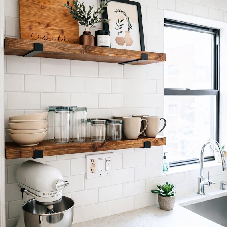 Two rich, medium-brown stained wood shelves mounted on a white tiled kitchen wall. The top shelf holds a decorative cutting board and framed art, while the bottom shelf is organized with kitchen essentials. Each shelf is supported by two sleek black metal brackets.
