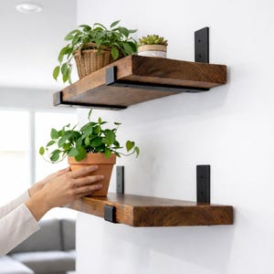 May include: Two rustic wooden shelves with black metal brackets are mounted on a white wall. The top shelf holds a wicker basket with a green plant and a small potted succulent. The bottom shelf has a terracotta pot with a green plant.