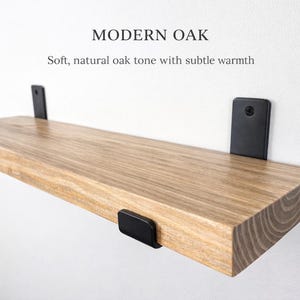 Peut inclure: &Eacute;tag&egrave;re flottante en ch&ecirc;ne clair avec des supports en m&eacute;tal noir, fix&eacute;e &agrave; un mur blanc. Le texte indique &laquo; MODERN OAK &raquo; et &laquo; Soft, natural oak tone with subtle warmth &raquo;. Design moderne.