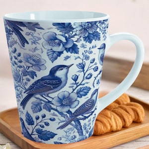 May include: A white ceramic mug with a blue floral and bird design. The mug has a curved handle and a wide rim. The design features blue birds, flowers, and foliage. The mug is sitting on a wooden tray.