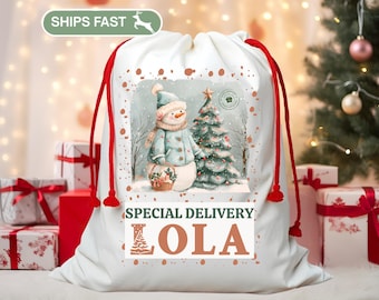 May include: White Christmas gift bag with red drawstring closure. The bag features a watercolor illustration of a snowman and Christmas tree. Text on the bag reads "Special Delivery Lola". The bag is surrounded by wrapped gifts.