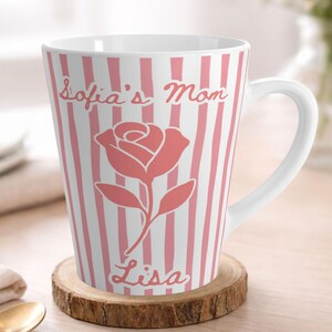 May include: A white latte mug with pink vertical stripes and a pink rose graphic. The mug has the text "Sofia's Mom Lisa" in a cursive font. The mug is on a wooden coaster.