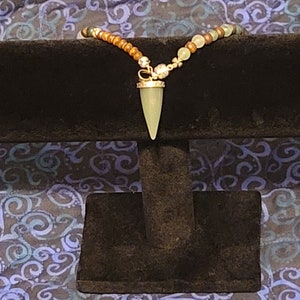 May include: A necklace with a light green stone pendant shaped like a point. The necklace is made of brown and green beads.
