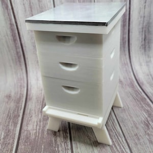 May include: A white, two-tiered beehive with a flat top and a base with legs. The hive has two drawers with curved handles and small, round ventilation holes. The hive is made of a solid, opaque material.