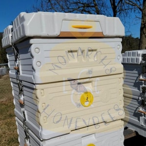 May include: A white beehive with a yellow and brown top. The beehive has a sign that says "Monarch Honeybees". There are bees flying around the bottom of the beehive.