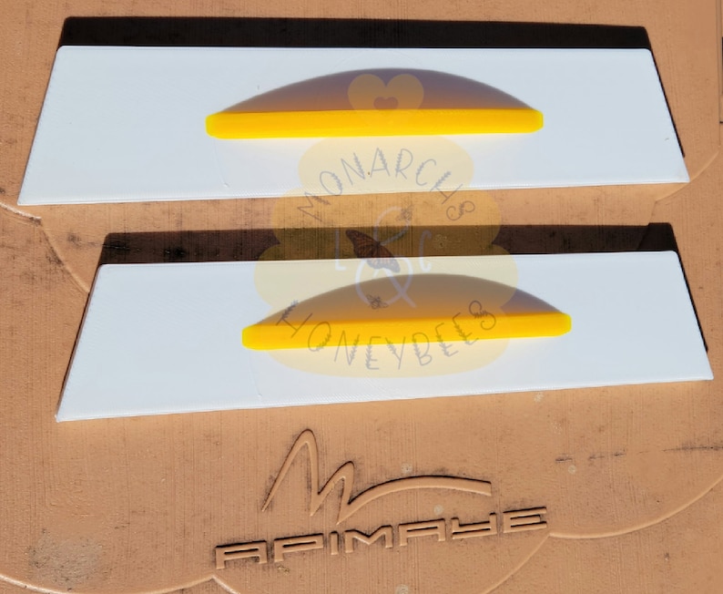 May include: Two white plastic beehive frames with yellow plastic inserts. The frames are labeled "Monarch's Honeybees".