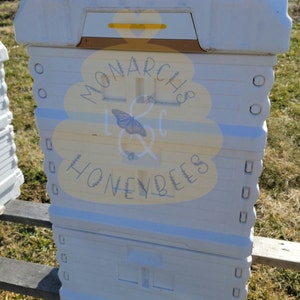 May include: White beehive with a yellow and brown top. The beehive has a label that says "Monarch & Honeybees".